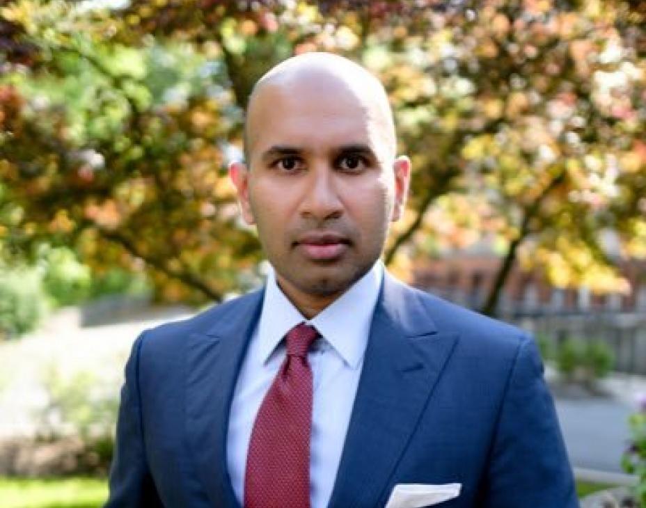 Dr. Davar in a blue suit facing forward with a rather serious demeanor. He is outdoors with autumn trees in the background.