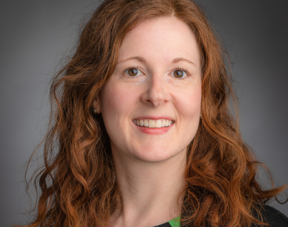 Dr. Erin Parry headshot. She is smiling facing forward. She has shoulder-length, wavy red hair, and is wearing a black jacket with patterned turquoise/blue blouse.