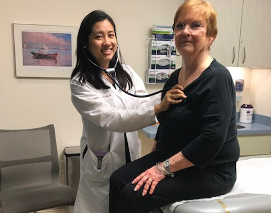 Dr. Jacqueline Garcia with a patient. Dr. Garcia is wearing a white coat and stethoscope, holding it up to the patient's heart. Both people are facing forward and smiling. They are located in a clinical room setting.