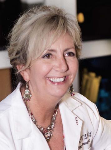 Dr. Gail Eckhardt smiling in a white coat, facing forward and positioned toward her right. She is wearing a necklace and dangly earrings. Hair is whitish blonde. Hopeful, cheerful look on her face.