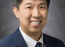 Headshot of Dr. Clinton Yam smiling and looking at the camera, in front of a dark gray backdrop.