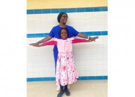 Dr. Kouya Francine standing behind Abigail, her patient. Both are outstretching their arms like wings and both are smiling. They appear to be in a clinical setting.