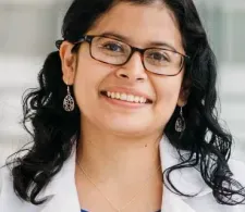 Dr. Jenny Ruiz wearing a white coat and smiling facing forward. She is wearing brown optical glasses, has black semi-straight and semi-curly hair, is wearing an azure blue blouse beneath her white coat, and has on dangly earrings.