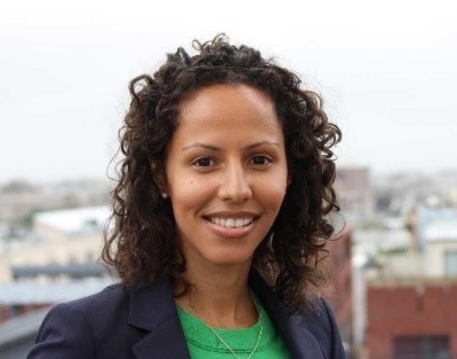 Headshot of Dr. Handy Marshall smiling. City skyline in background. Wearing light green blouse with navy blue blazer. Brown, curly hair.