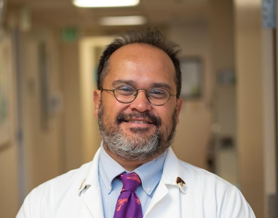 Dr. Flavio Rocha facing forward and smiling. He is wearing a white coat. He's in a well-lit hallway that appears to be in a hospital or clinic.