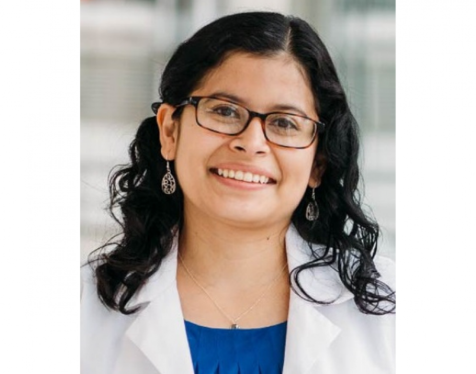 Dr. Jenny Ruiz wearing a white coat and smiling facing forward. She is wearing brown optical glasses, has black semi-straight and semi-curly hair, is wearing an azure blue blouse beneath her white coat, and has on dangly earrings.
