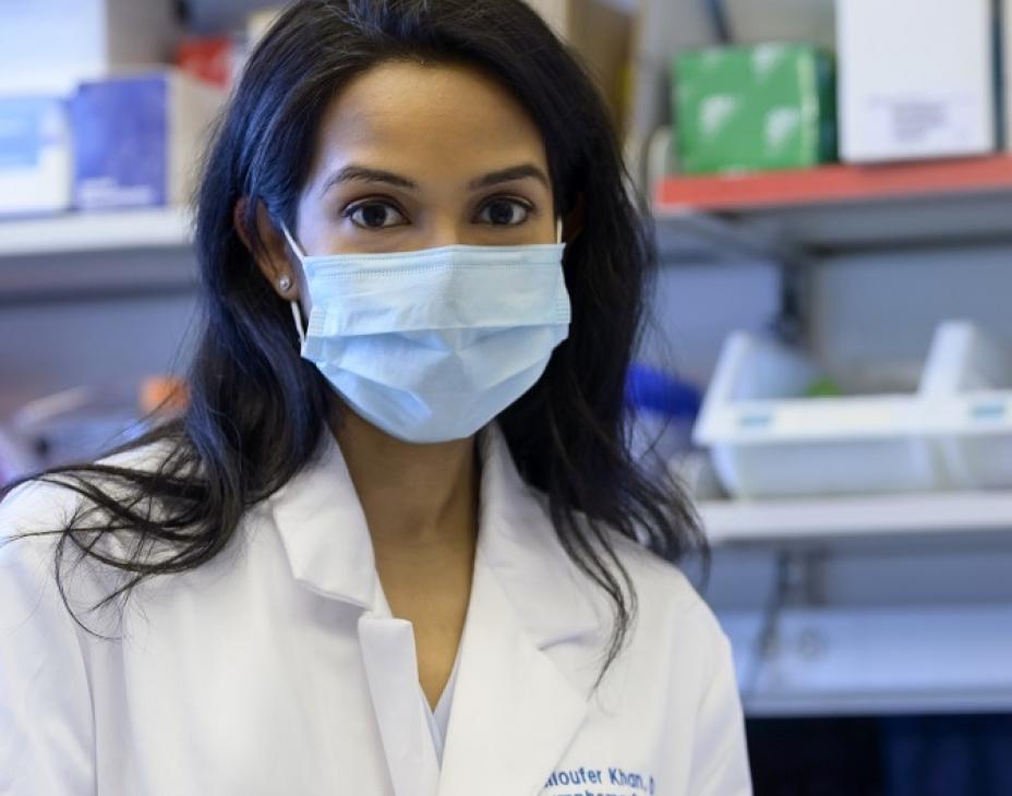 Niloufer Khan, wearing a medical white coat and a mask, facing forward. She is in a clinical laboratory settings, holding a document.