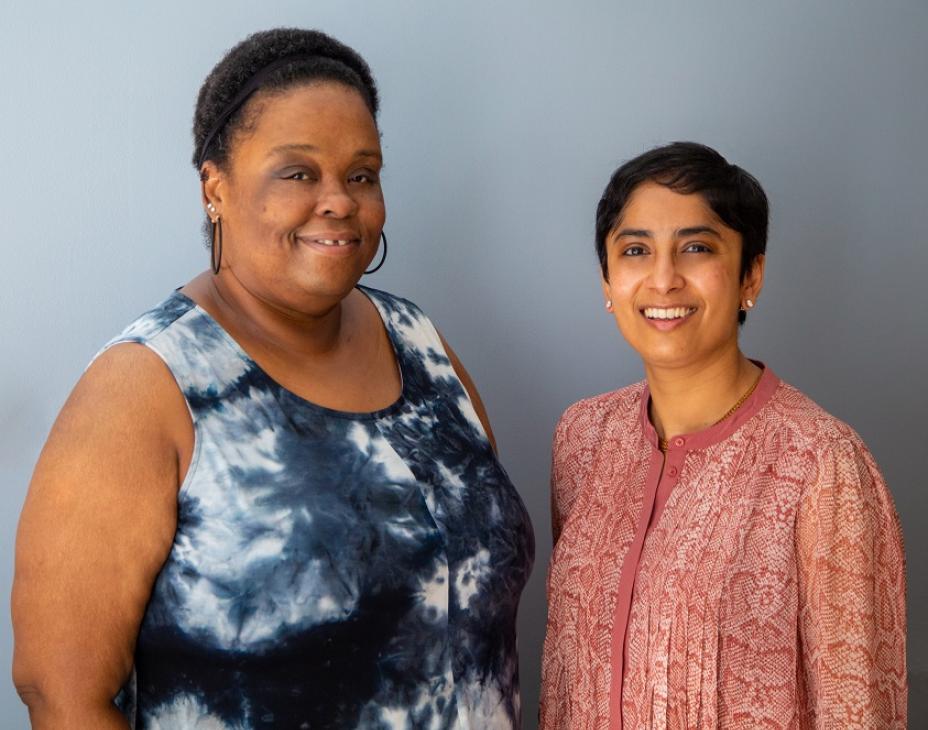 From left to right: Menekia, a patient with T-cell lymphoma, smiling facing forward and wearing a blue tie-dye shirt. On the right, her doctor, Salvia Jain, MD, is also smiling, and is wearing a pinkish-red shirt.