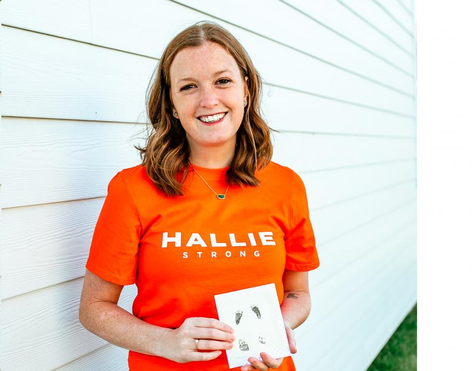 Bethany Hart holding a photo and wearing an orange shirt and blue jeans. She has red shoulder-length hair and is standing in front of a white wall.