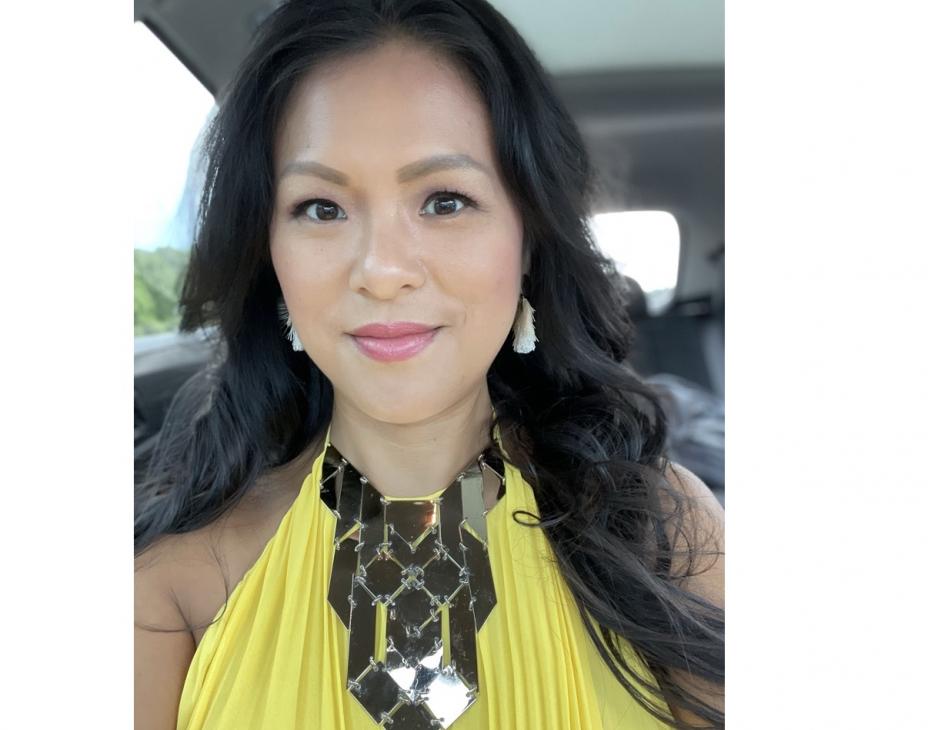 Selfie of Mai Achong. She is seated in a car, wearing a yellow dress and a gold platelet necklace. Her hair is black and shoulder-length. She is smiling facing forward.