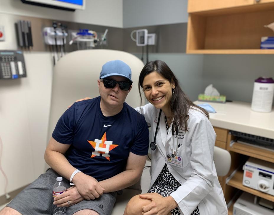 Dr. Ferrarotto with a patient in clinic. Dr. Ferrarotto is wearing a white coat and stethoscope, and is smiling facing forward. She is next to her patient, who is wearing black sunglasses and a dark blue shirt.