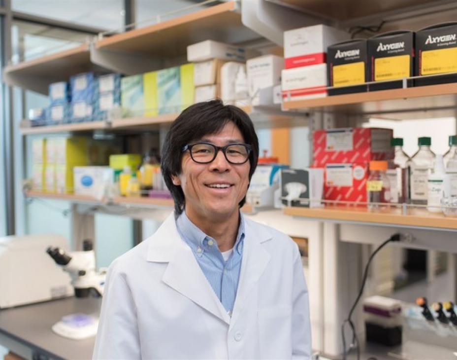 Dr. William Matsui, wearing a white coat and glasses, and is located in a laboratory. He has black hair and is smiling facing forward.