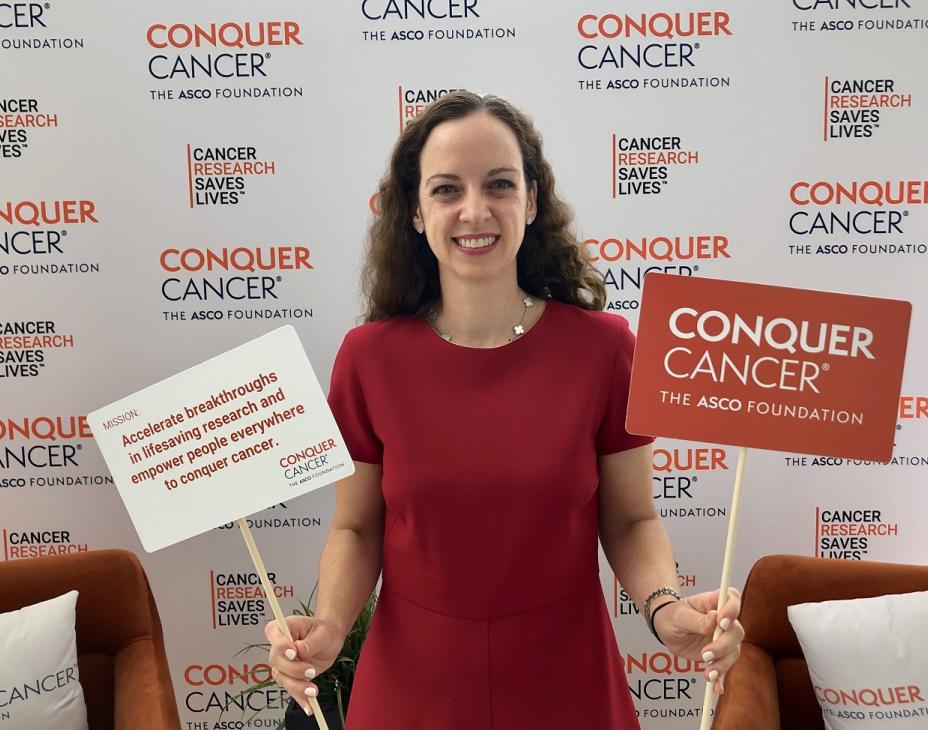 Dr. Lauren Byers in front of a patterned background that read "Conquer Cancer, the ASCO Foundation," and "Cancer Research Saves Lives." She is wearing a red dress and has shoulder-length brown hair, and is holding two signs. One contains the Conquer Cancer logo. The other cannot be easily interpreted,