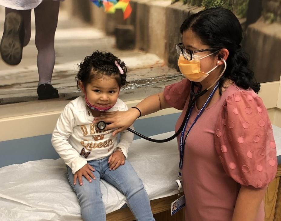 Dr. Jenny Ruiz caring for a pediatric patient. She is holding a stethoscope to the child patient's heart. The patient is smiling facing forward.