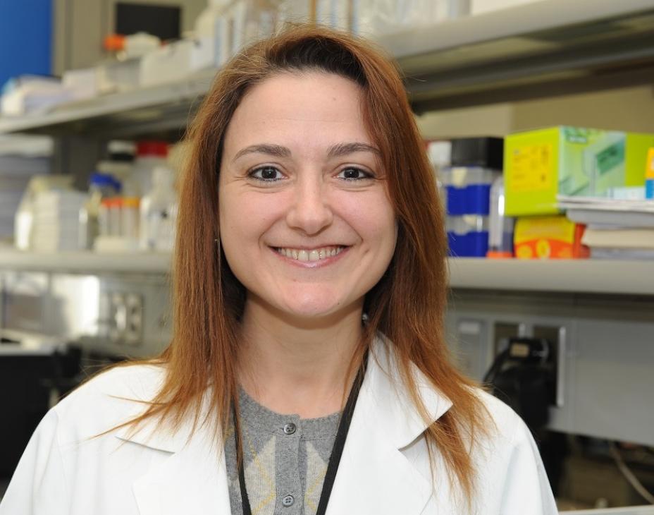 Headshot of Dr. Raya Saab in a laboratory setting. She is wearing a white coat and has shoulder-length red-brown hair. She is smiling facing forward.