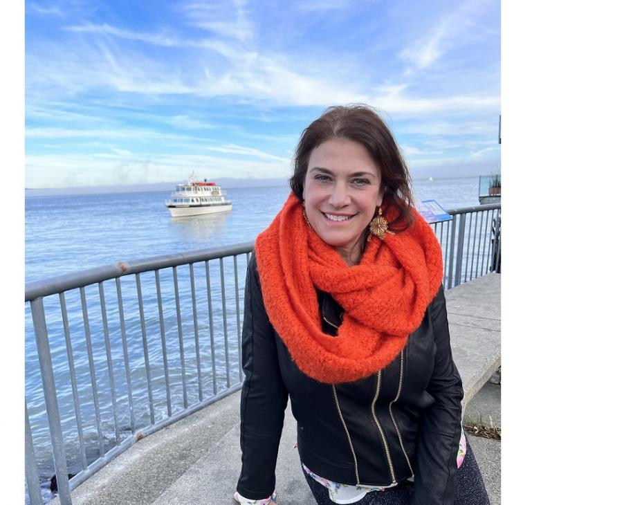 Lillian Kreppel wearing an orange scarf and a black long-sleeved outfit. She is smiling facing forward, sitting on a ledge nearby a bay. The sea and sky merge on the horizon and it is sunny and blue.