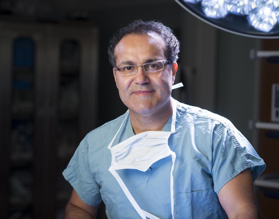 Headshot of Dr. Alfredo Quiñones-Hinojosa in surgical/medical scrubs. He's wearing glasses and has a white mask hanging on his chest, and he's standing in a surgery room.