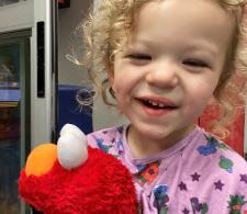 Leo with medium-length curly hair, smiling and laughing, while holding an Elmo plushie.