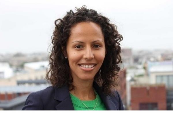 Dr. Handy Marshall headshot, smiling at the camera. City skyline in the background, bright gray sky. Wearing a bright green blouse with navy blue blazer. Brown, curly hair.