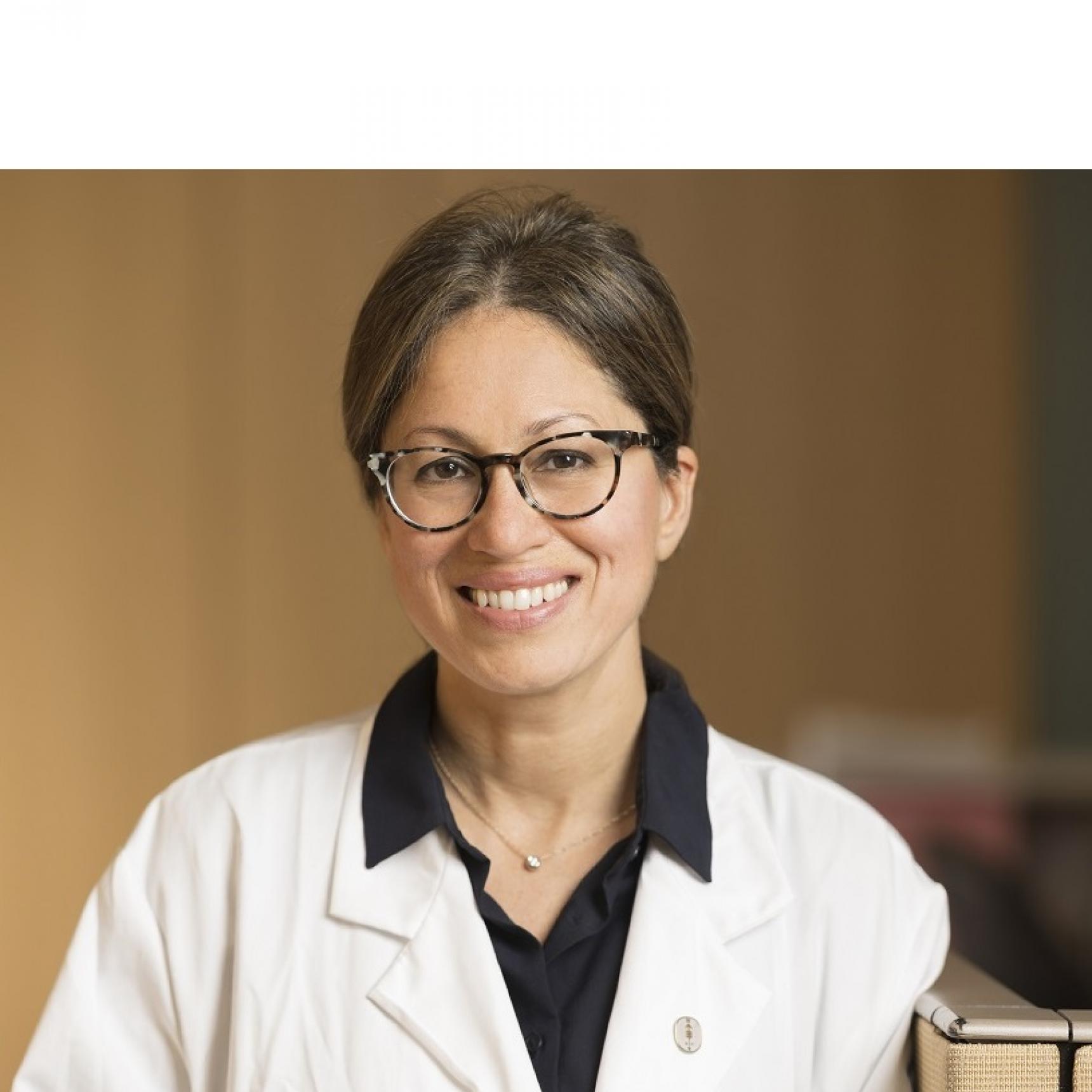 Headshot of Dr. Shanu Modi in a clinical sitting wearing a white coat, glasses, and brown hair. She is smiling facing forward.
