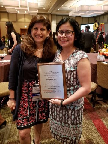 Dr. Jenny Ruiz is standing to the left of Dr. Julia Glade Bender, her ASCO mentor. Dr. Ruiz is wearing a black and white patterned dress and is holding her 2018 RTA award plaque, while smiling at the camera. Dr. Glade Bender is next to Dr. Ruiz, wearing a black blouse and dark maroon skirt.