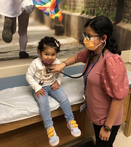 Dr. Jenny Ruiz with a young child patient. The patient appears to be about 3-4 years of age. Dr. Ruiz is wearing a mask and is using a stethoscope on her patient's chest. The patient is gently smiling and facing downward, appearing happy to be in Dr. Ruiz's care.