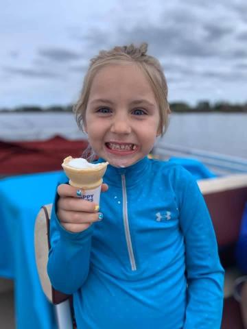 Kenedi Schoeneck, wearing a blue jacket and holding a vanilla ice-cream cone. She is smiling widely, while located on a boat. The background consists of a body of water and a forested horizon.