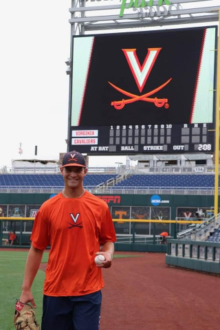 Jake Berry wearing an orange shirt and a baseball cap, with a baseball mitt. He is standing on a baseball field at the University of Virginia.
