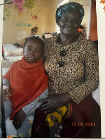 Abigail and her grandmother, Suzan. Abigail is a young child and is wearing a red shirt. Suzan is holding Abigail, and is wearing a light-brown shirt. Both are facing forward and appear to be in a clinical environment.