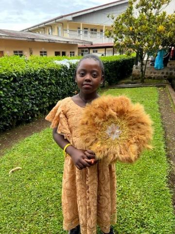 Abigail wearing a gold dress with a large golden flower by her shoulder. She is standing outside in a green yard and is facing forward.