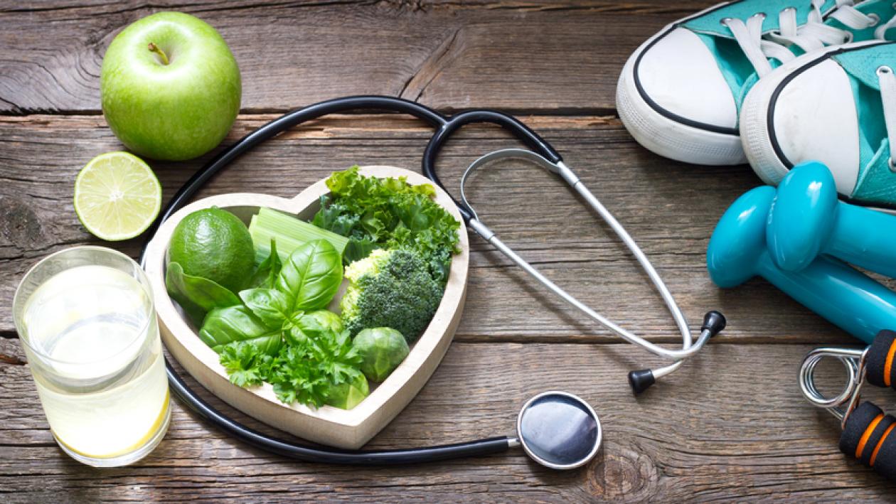 An image with a heart-shaped bowl with green vegetables, a doctor's stethoscope, pair of running shoes, hand weights, a glass of water, and a green apple
