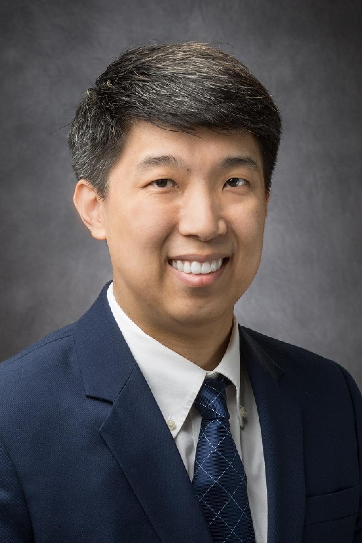 Headshot of Dr. Clinton Yam smiling and looking at the camera, in front of a dark gray backdrop.