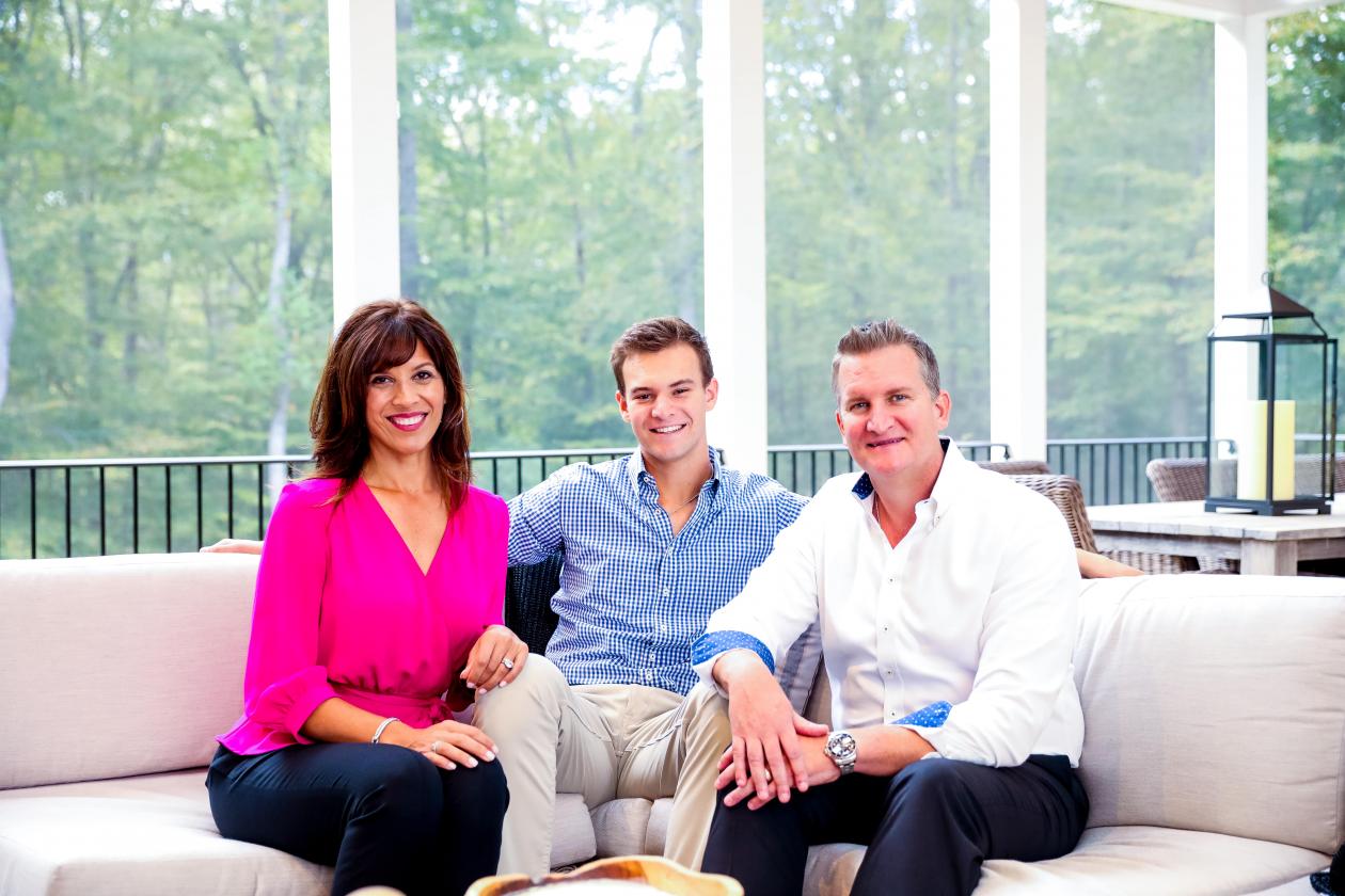 Left to right: Nancy Berry, Jake Berry, Alex Berry. All three are seated facing forward and smiling, located in a screened-in sun-room with white columns and green trees visible in the background. Nancy is wearing a bright pink shirt with dark blue pants. Jake is wearing a light blue shirt with khaki plants. Alex is wearing a white shirt with black pants.