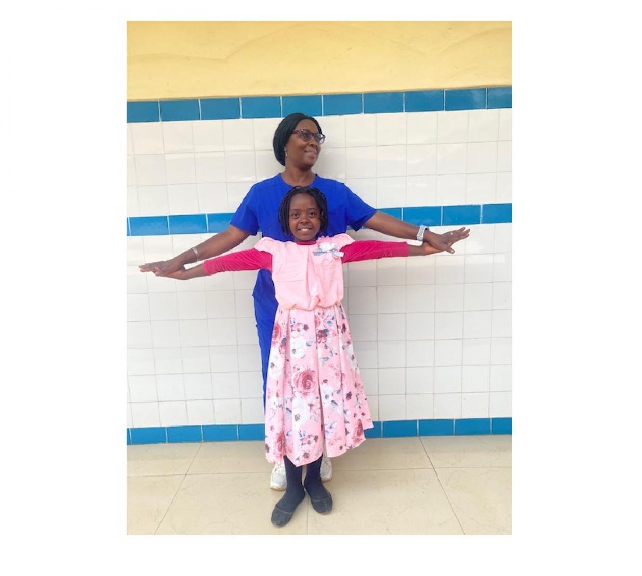 Dr. Kouya Francine standing behind Abigail, her patient. Both are outstretching their arms like wings and both are smiling. They appear to be in a clinical setting.