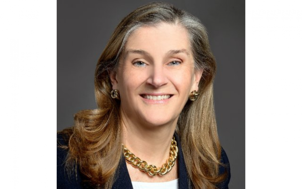 Headshot of Conquer Cancer CEO Nancy Daly. She has shoulder-length brown hair and is wearing a gold necklace, white shirt and black blazer, smiling facing forward against a gray background.
