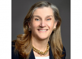 Headshot of Conquer Cancer CEO Nancy Daly. She has shoulder-length brown hair and is wearing a gold necklace, white shirt and black blazer, smiling facing forward against a gray background.