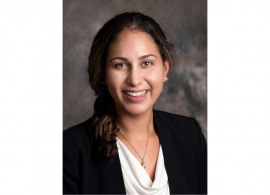 Headshot of Dr. Eleanor Taranto. She is smiling facing forward against a gray background and has shoulder-length brown hair, and is wearing a black blazer over a white blouse with a necklace.