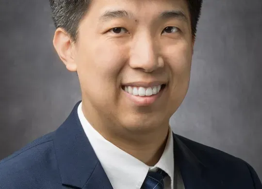 Headshot of Dr. Clinton Yam smiling and looking at the camera, in front of a dark gray backdrop.