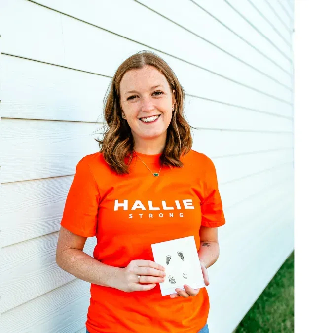 Bethany Hart holding a photo and wearing an orange shirt and blue jeans. She has red shoulder-length hair and is standing in front of a white wall.