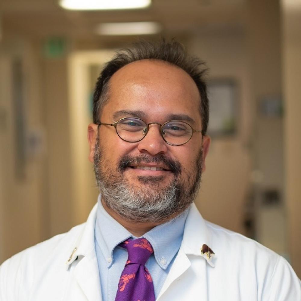 Dr. Flavio Rocha facing forward and smiling. He is wearing a white coat. He's in a well-lit hallway that appears to be in a hospital or clinic.