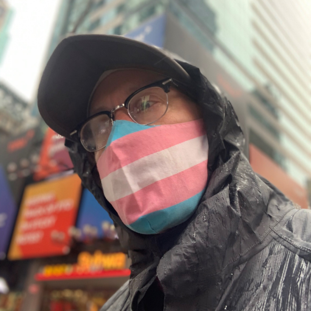 Scout, wearing a transgender pride flag (consisting of blue, pink, and white stripes). He is wearing a black rain jacket and has a baseball cap on. He appears to be standing in an urban environment, with skyscrapers in the background.