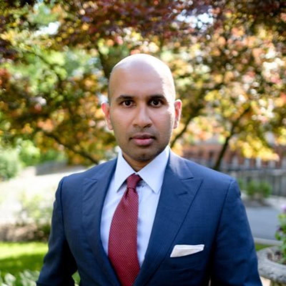 Dr. Davar in a blue suit facing forward with a rather serious demeanor. He is outdoors with autumn trees in the background.