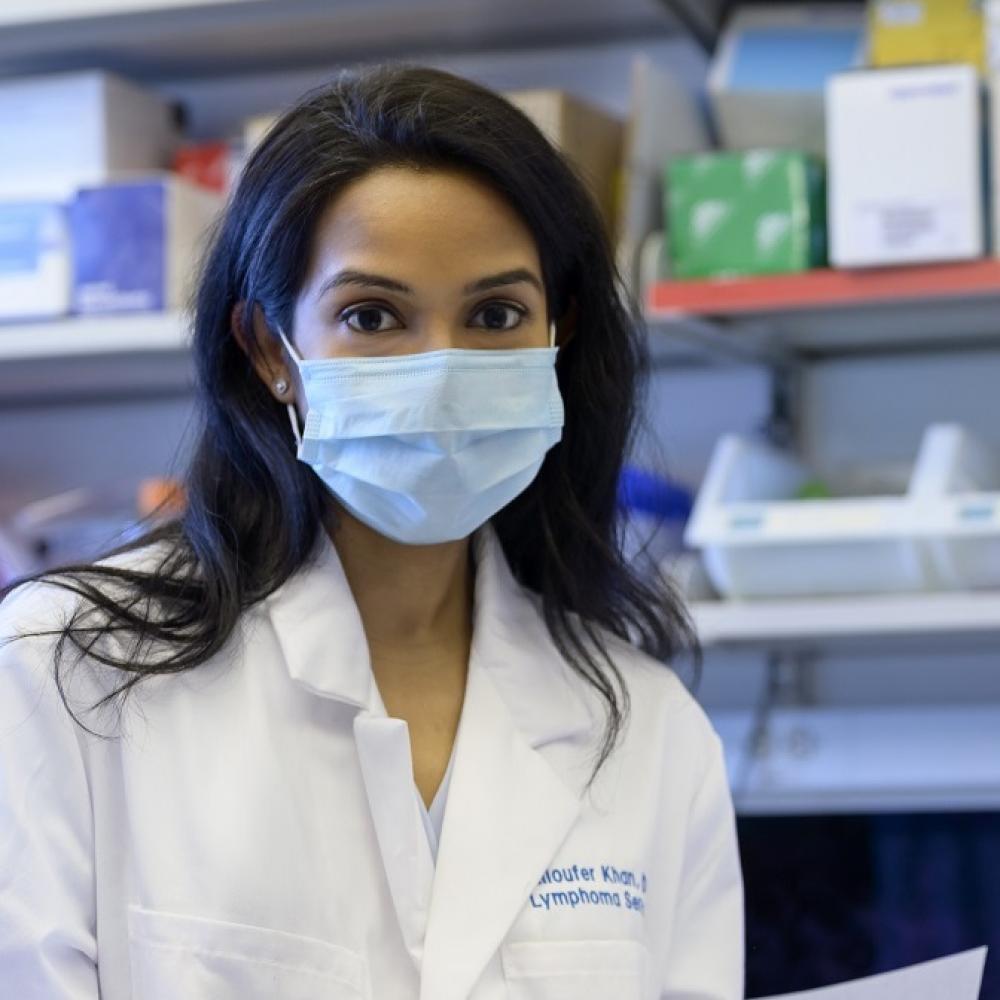 Niloufer Khan, wearing a medical white coat and a mask, facing forward. She is in a clinical laboratory settings, holding a document.