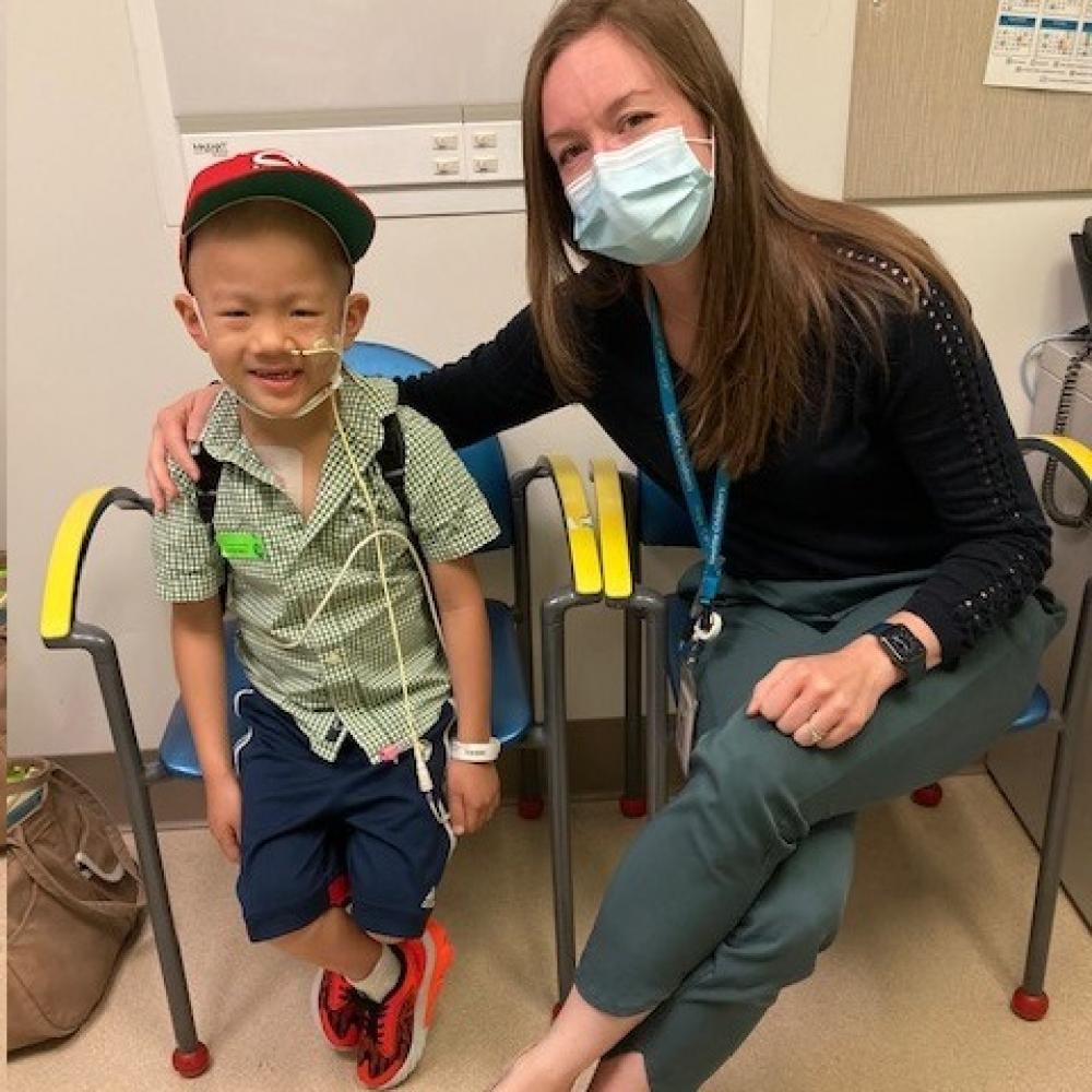 Dr. Molly Taylor sitting with a patient. Dr. Taylor is wearing a mask, black shirt, and jeans. Her patient appears to be very young, is wearing a green shirt, blue shorts, and a red cap. Both are smiling and facing forward.