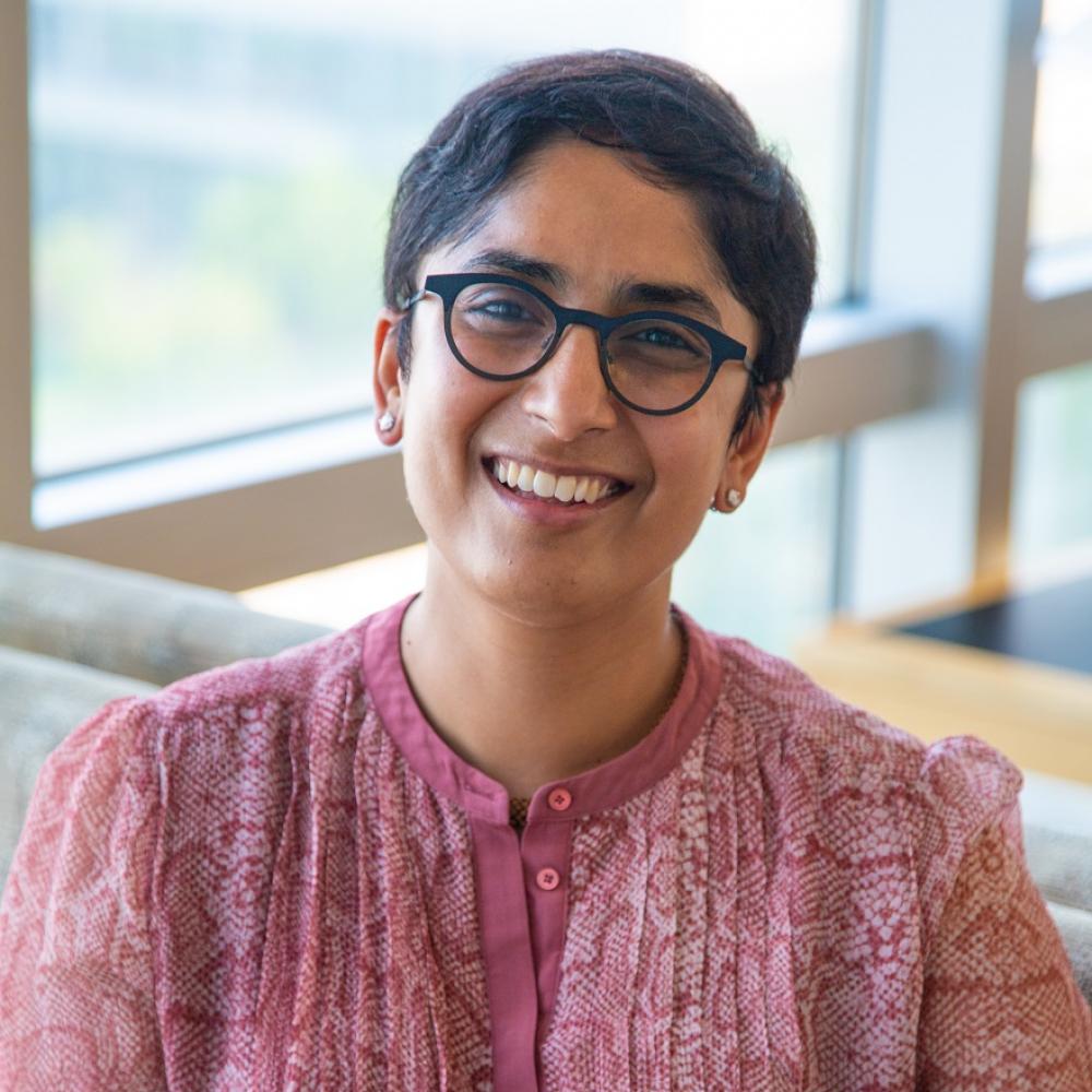 Dr. Salvia Jain smiling facing forward. She's sitting in a bright room with natural light. She's wearing a light pinkish-red shirt and reading glasses, and has short black hair.