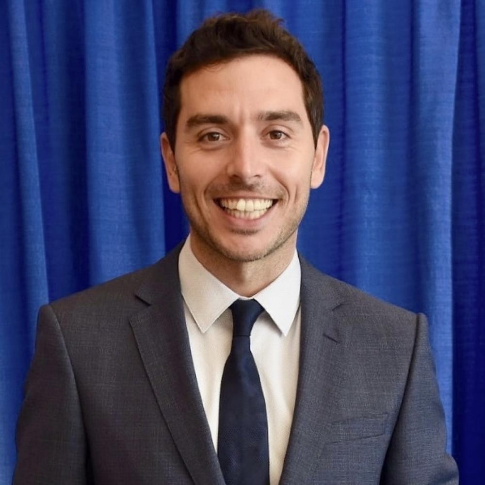 Headshot of Dr. Marc Oliva smiling facing forward. He is wearing a dark-gray suit and has short dark-brown hair. He is in front of a blue curtain background.