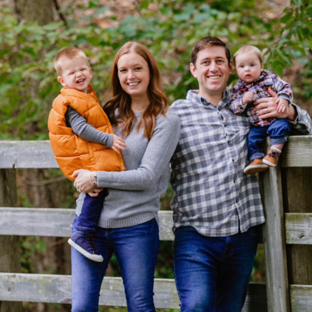 Bethany Hart pictured next to her husband and two sons. They are standing by a fence outdoors and smiling facing forward.