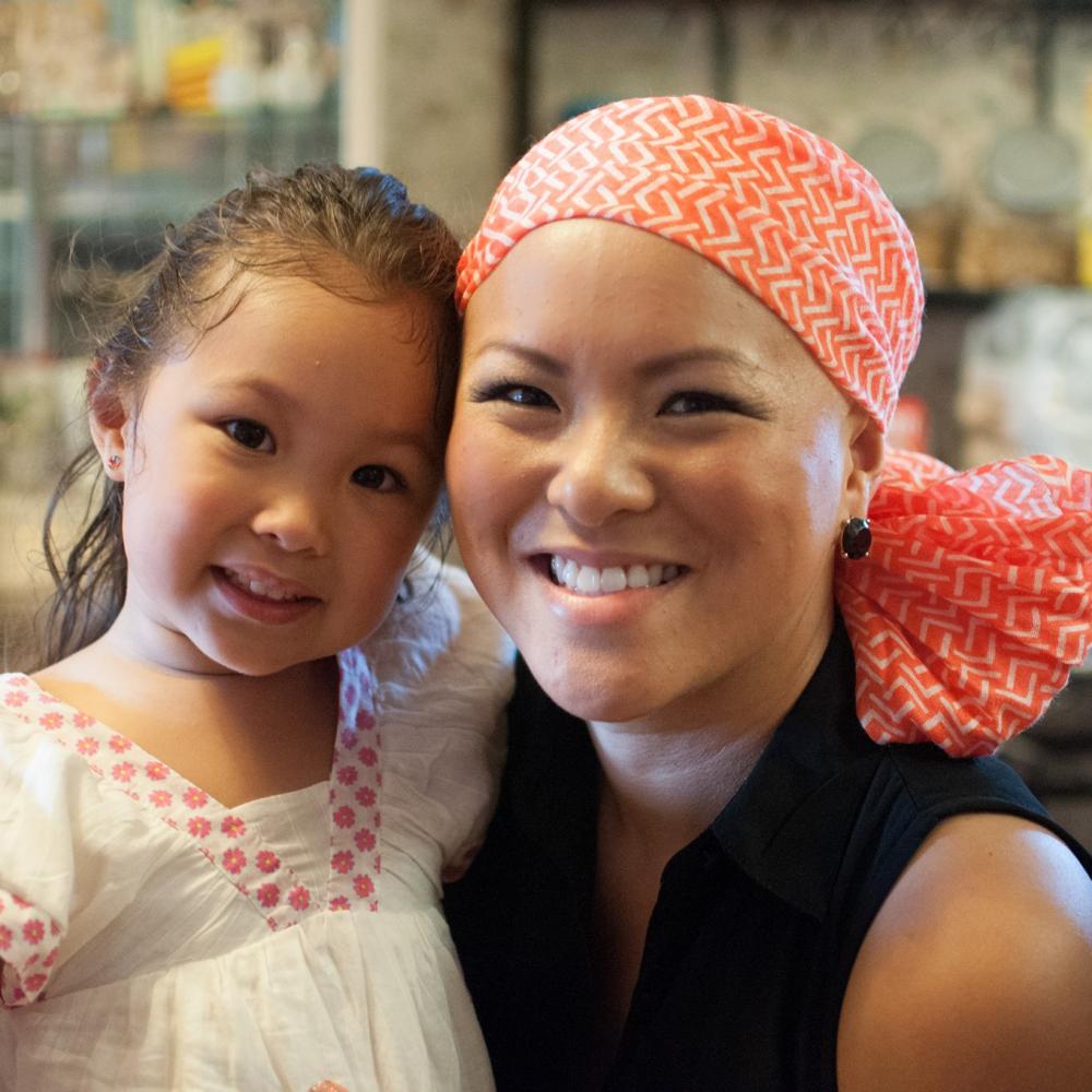 Mai Achong with her daughter, Angelina. Mai is on the right, with an orange bandana on her head and wearing a black blouse. Angelina is on the left, wearing a white shirt and leaning on Mai. Both are smiling facing forward.