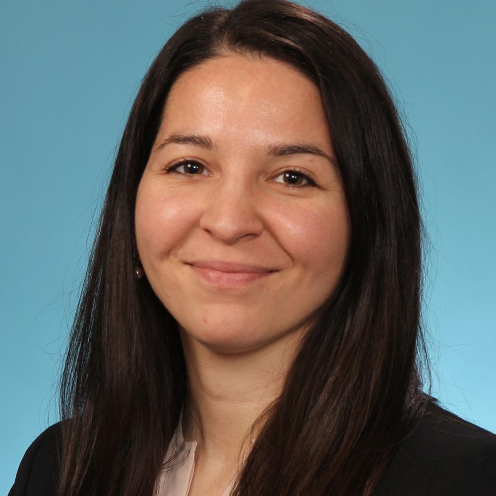 Headshot of Dr. Stephanie Markovina. She has long dark-brown hair and is wearing a black blazer with tan blouse, against a light-blue background. She is smiling facing forward.