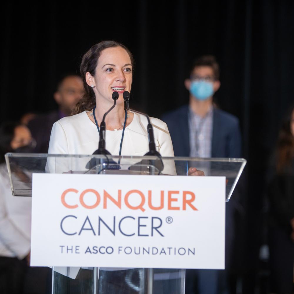 Dr. Lauren Byers speaking at the 2022 ASCO Annual Meeting. She is wearing a white outfit and is standing at a podium, announcing awards during the Grants & Awards ceremony. Recipients can be seen standing in the background. Dr. Byers is smiling and speaking.